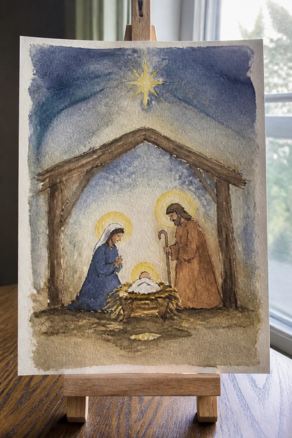 Watercolor nativity scene with Mary in blue, Joseph with staff, and haloed baby in manger under star.