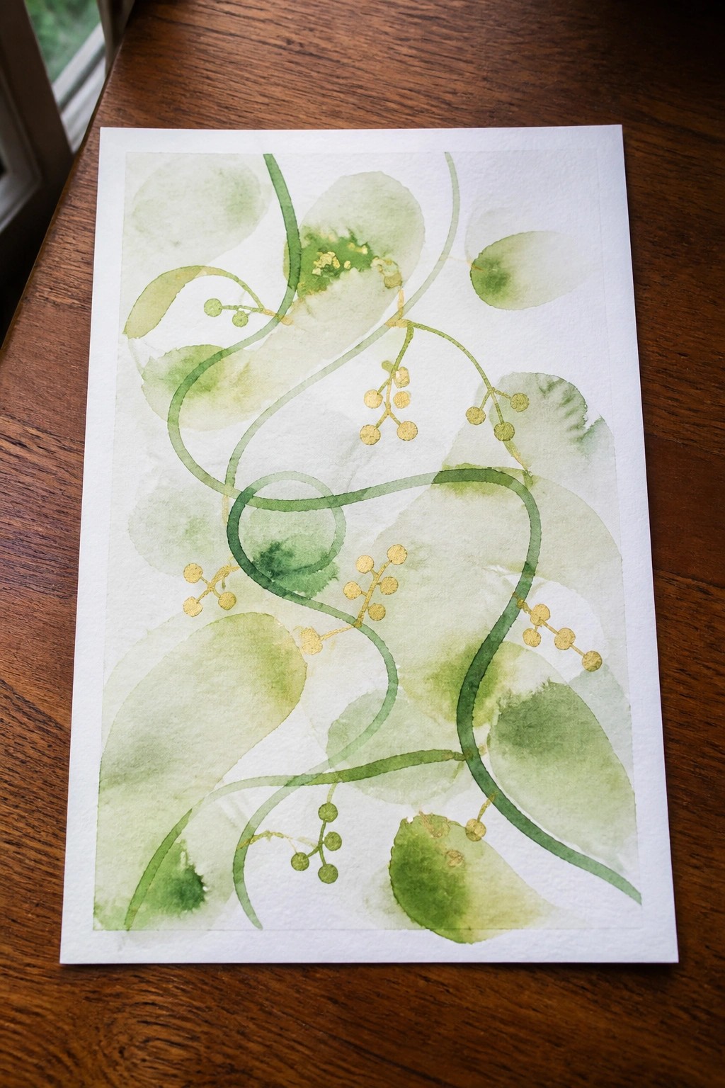 Abstract green watercolor leaves, vines, and gold berry clusters on white paper over wood.