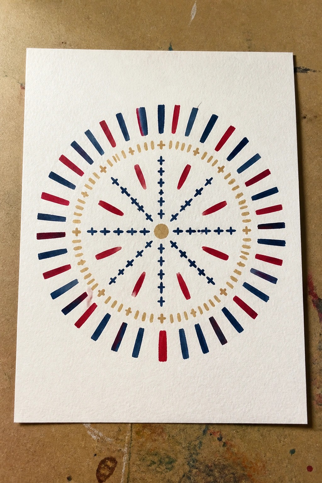 Watercolor mandala with red blue rays, gold crosses, dashes, and central dot on paper