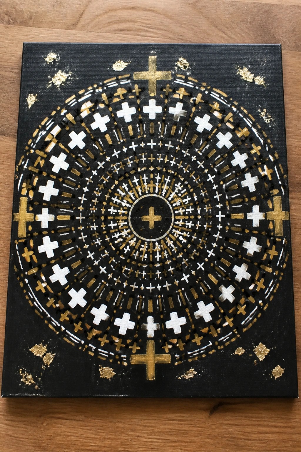 Black canvas mandala of concentric gold and white crosses with scattered gold leaf flakes