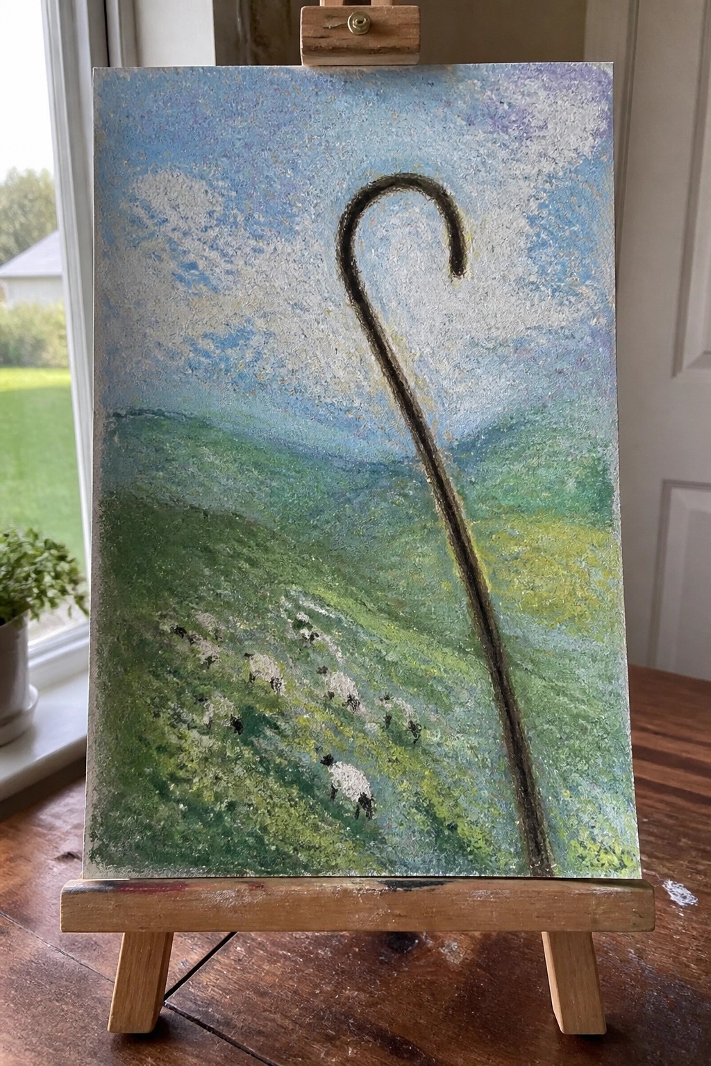 Painting on easel shows green hills, sheep flock, and tall black shepherd's crook under blue sky.