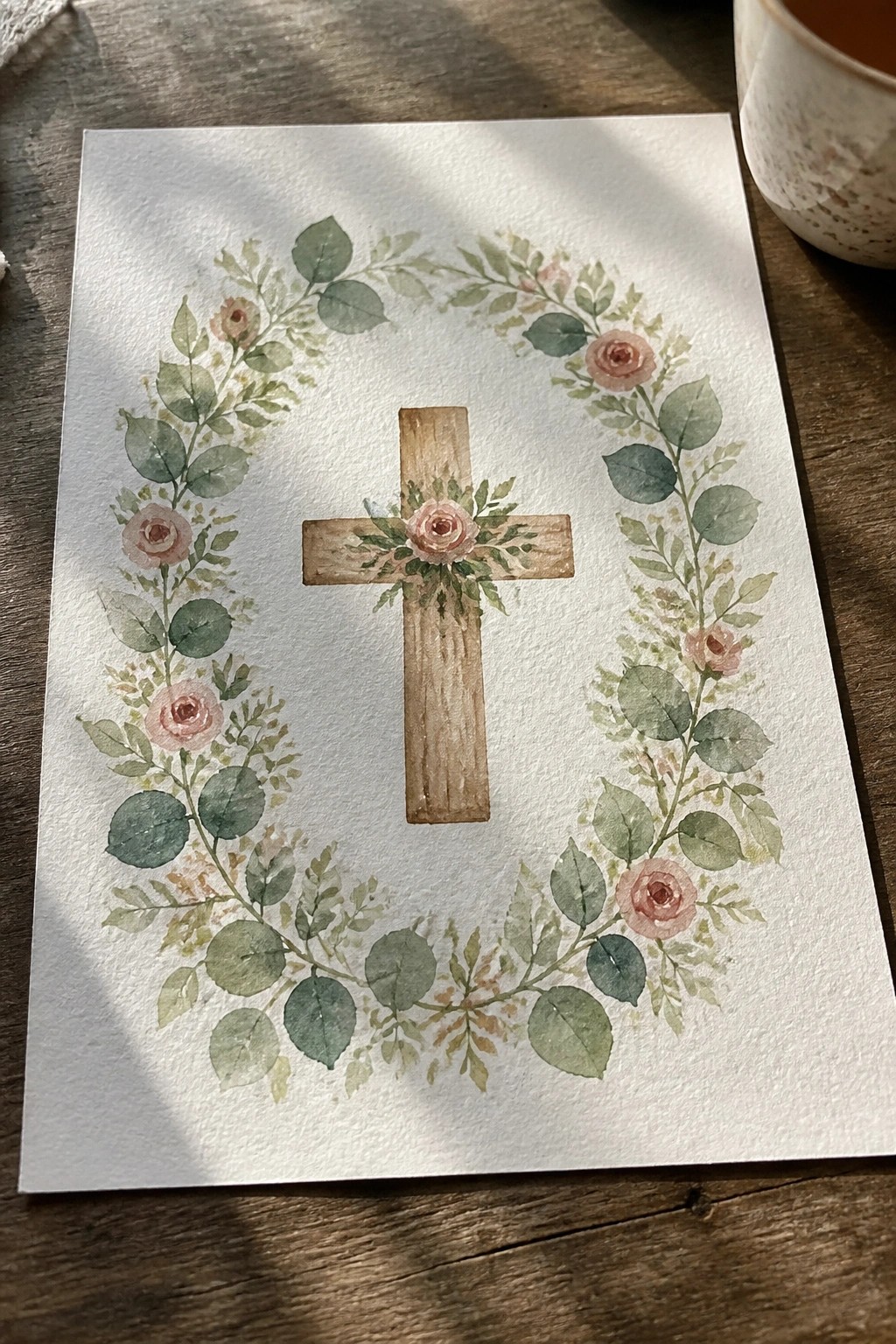 Watercolor cross centered in circular wreath of pink roses and green leaves on paper