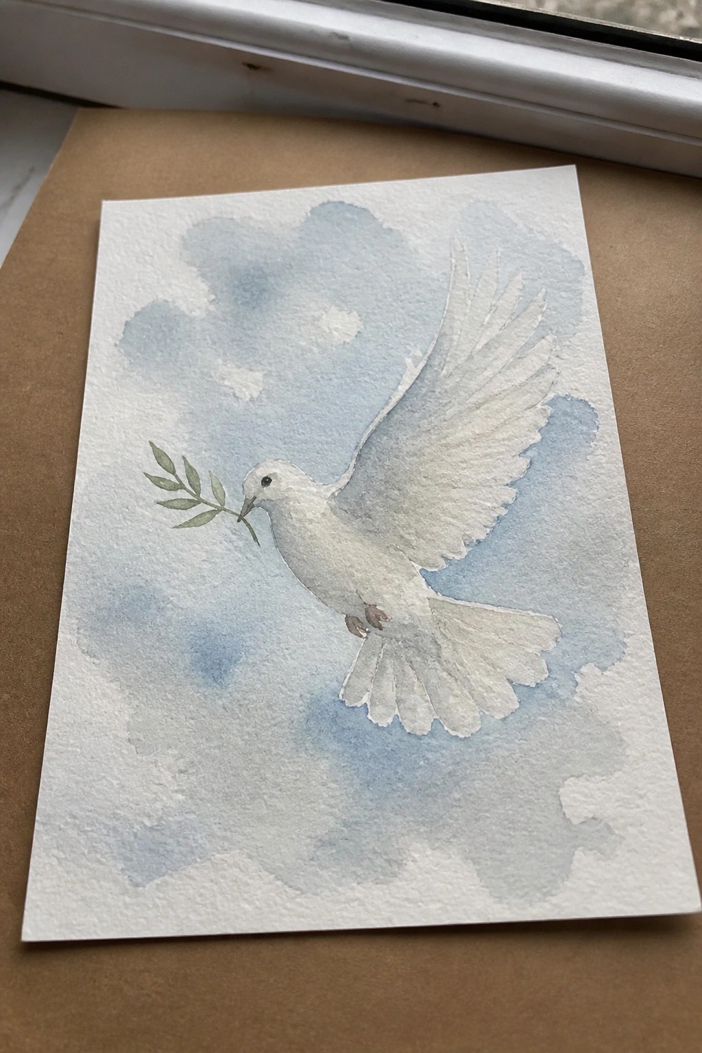 Watercolor painting of white dove with olive branch flying through cloudy blue sky