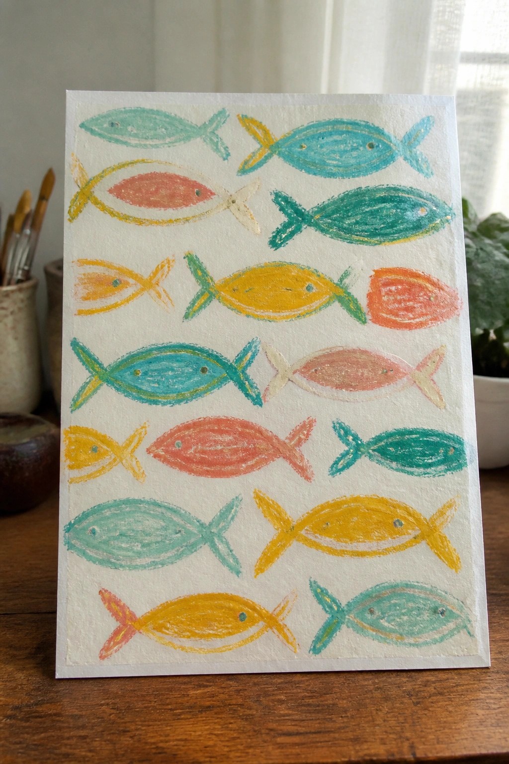 A white sheet of paper displays rows of colorful fish painted in teal, yellow, orange, and red on a wooden surface.