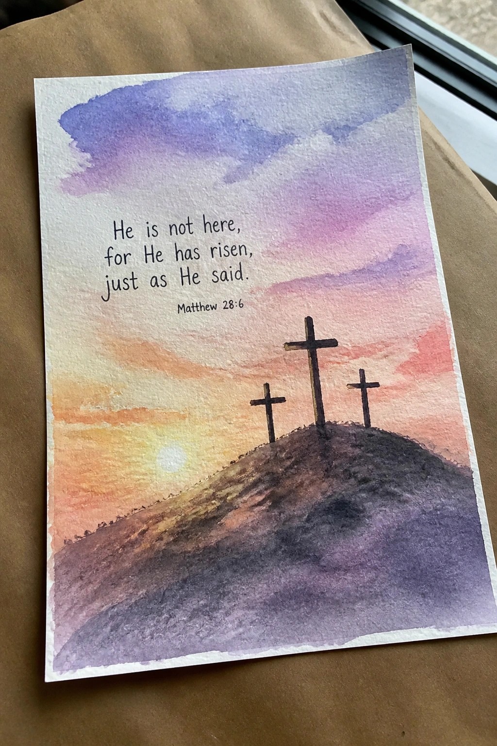 Watercolor painting of three crosses on a hill under vibrant purple-pink sunset sky with scripture text