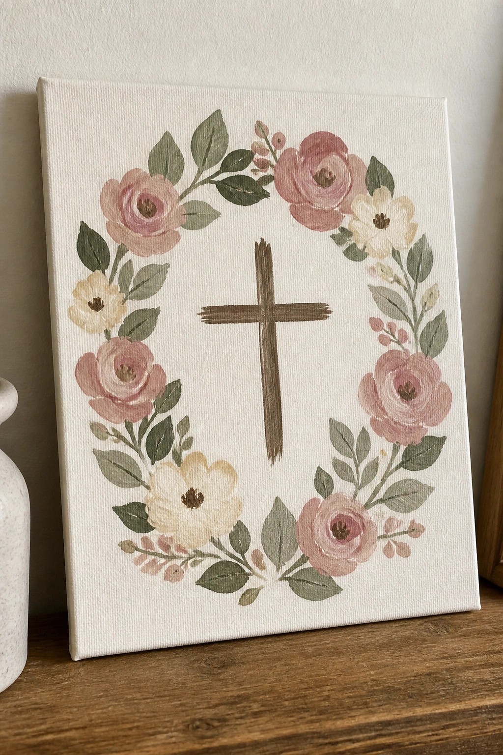 Floral wreath of pink roses and green leaves encircling brown cross on white canvas