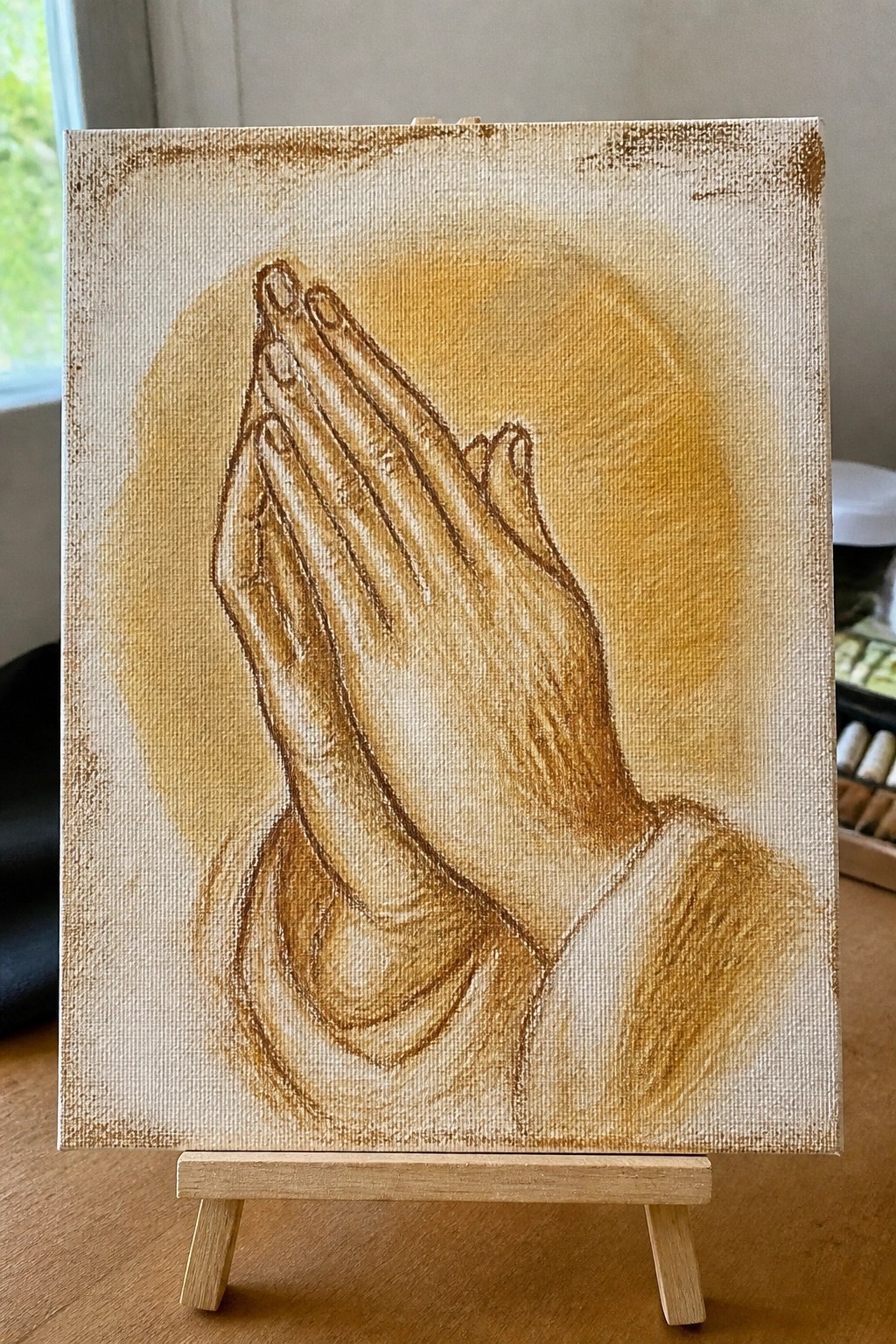 A canvas painting of praying hands with a soft brown glow behind them.