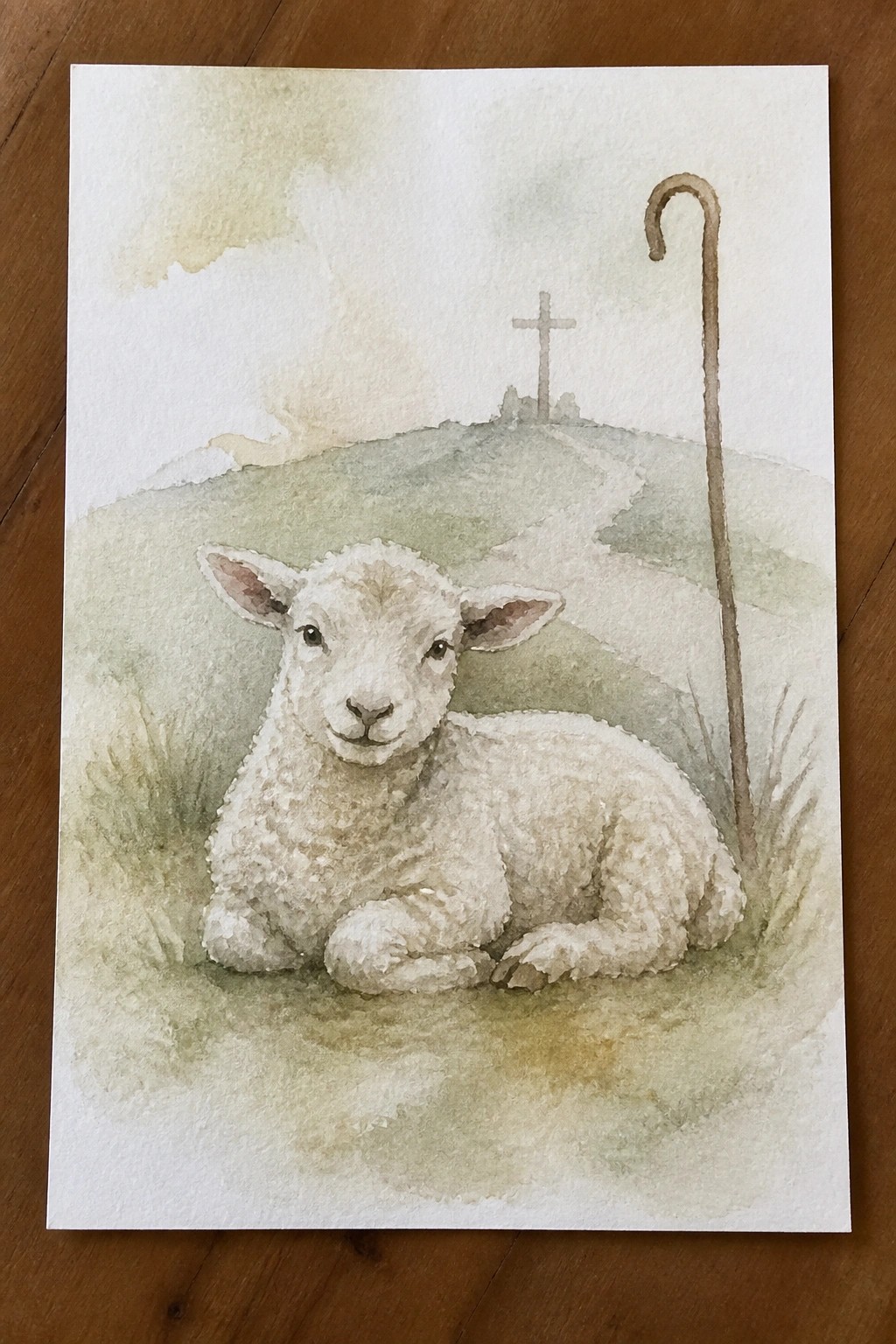 Watercolor of white lamb resting on grass beside shepherd's crook with cross on distant hill