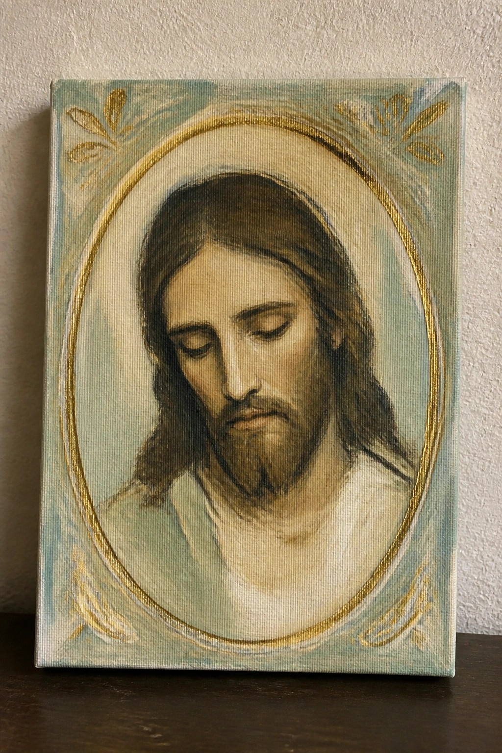 Painting of Jesus with bowed head and closed eyes in ornate gold oval frame on teal.
