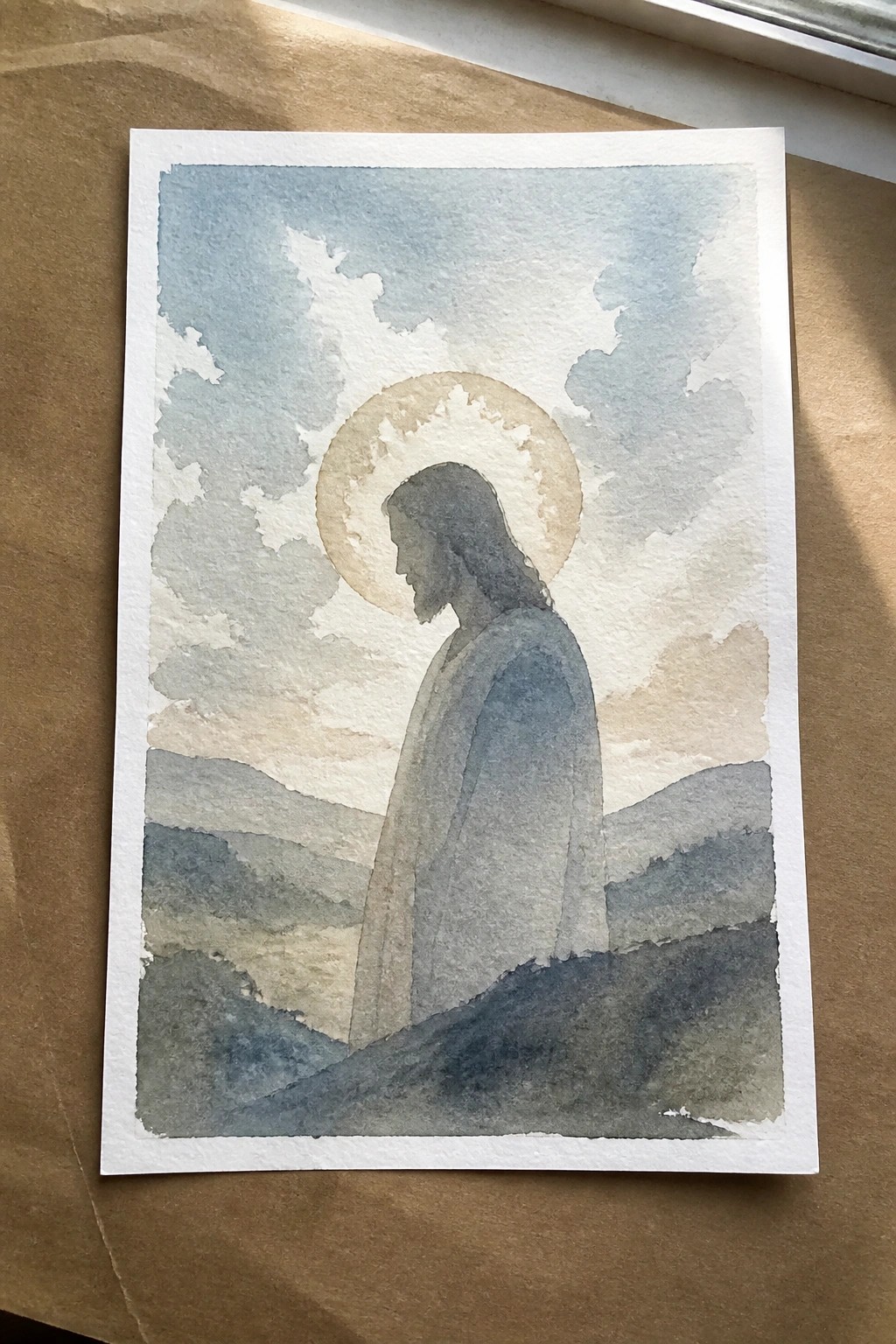 Watercolor of Jesus in gray robe with golden halo before cloudy blue mountains.