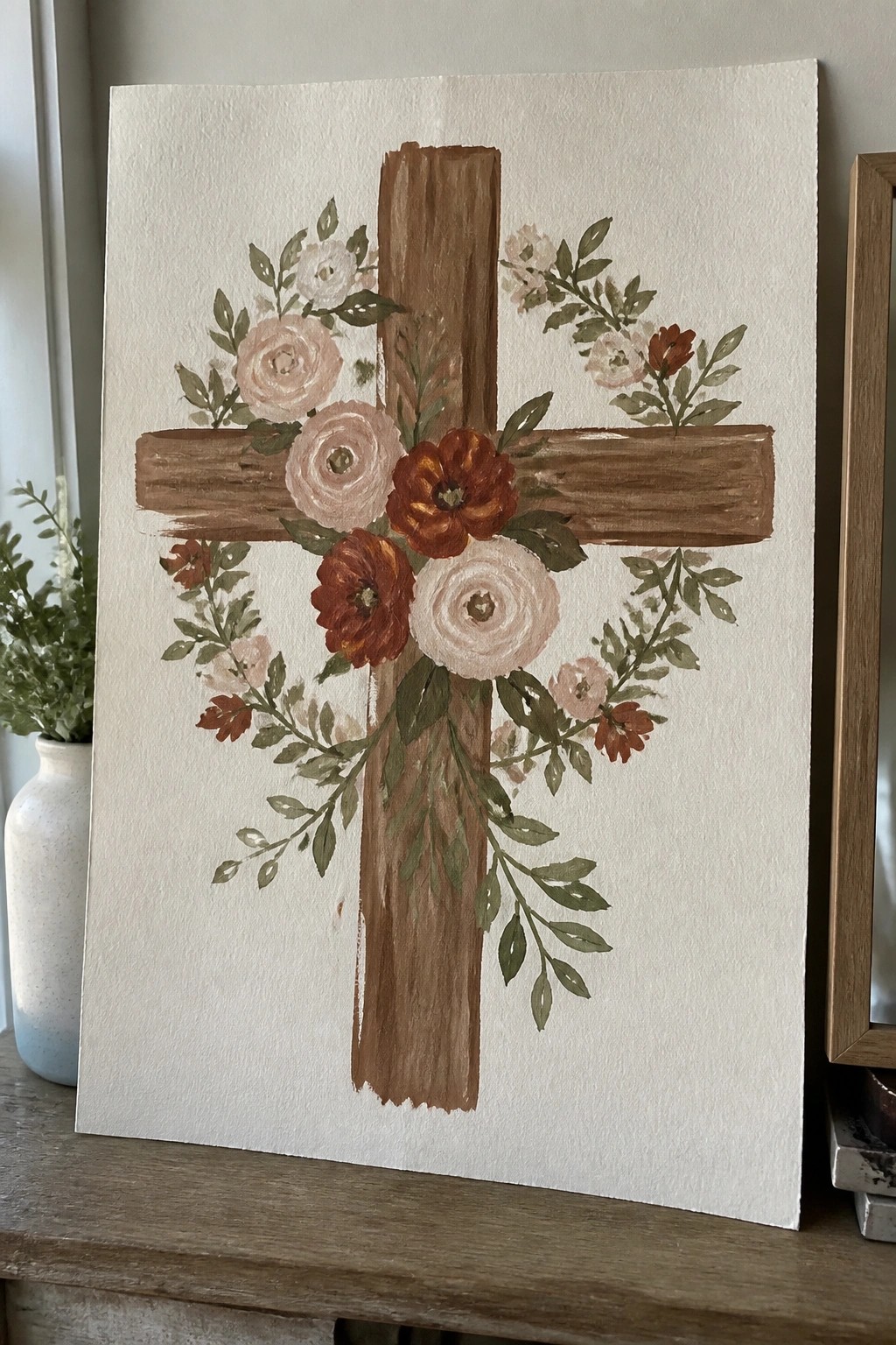 A painting of a brown wooden cross with pink, white, and rust flowers plus green leaves arranged around the center on white paper.