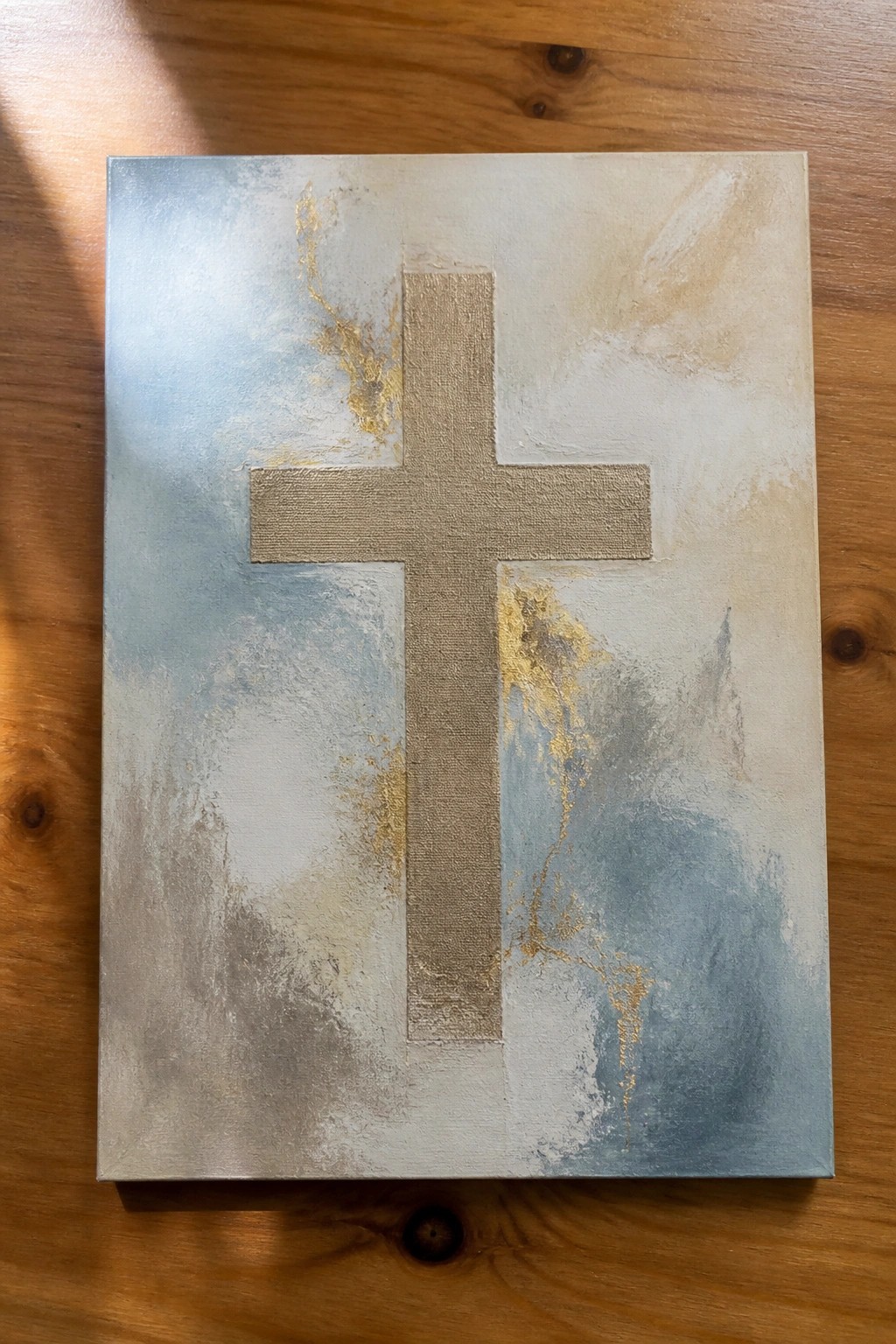 Abstract painting with textured beige cross on blue, gray, and gold background atop wood