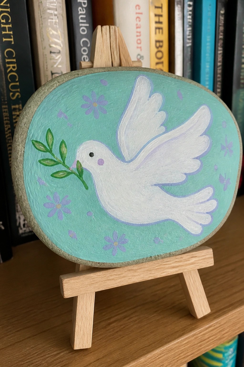 Turquoise painted rock with white dove holding olive branch amid purple flowers on easel