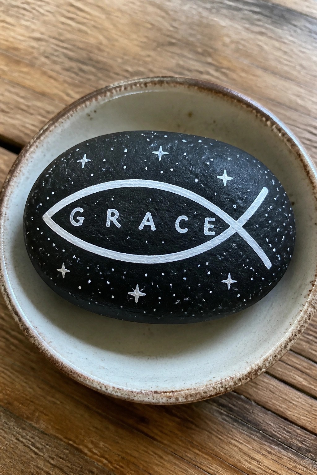 Black stone with white fish symbol spelling GRACE amid stars in a ceramic bowl on wood