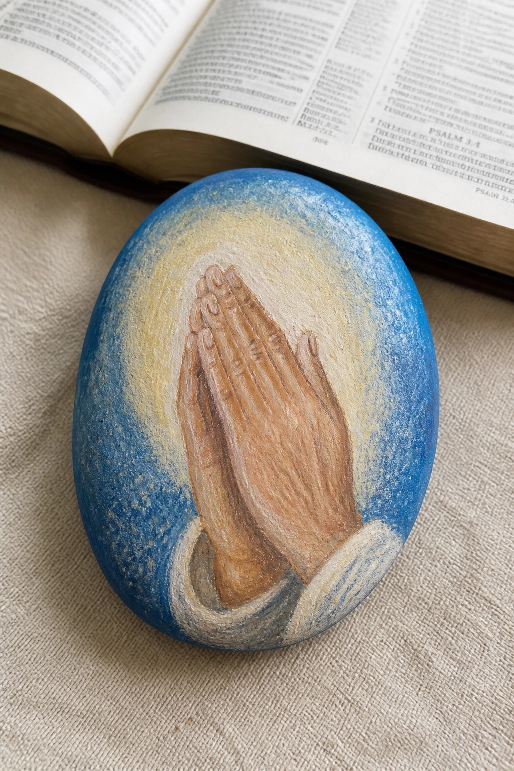 Painted blue rock with praying hands, yellow glow, and open Bible behind