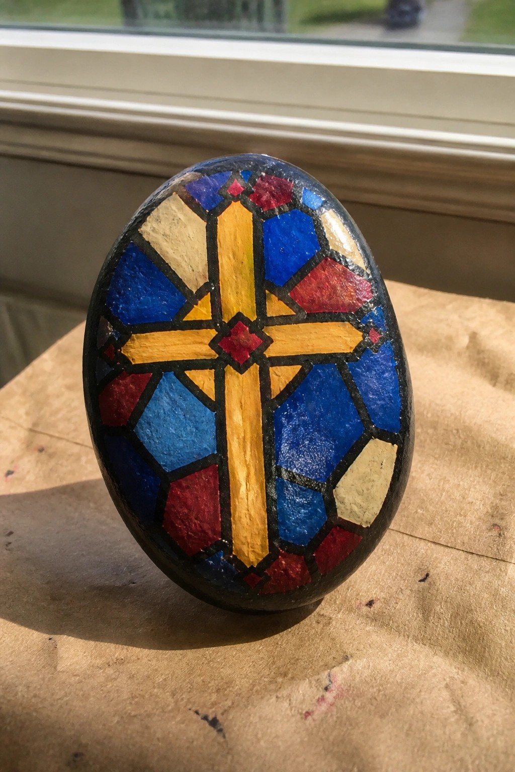Painted oval rock with yellow cross in blue, red, and beige stained-glass pattern.