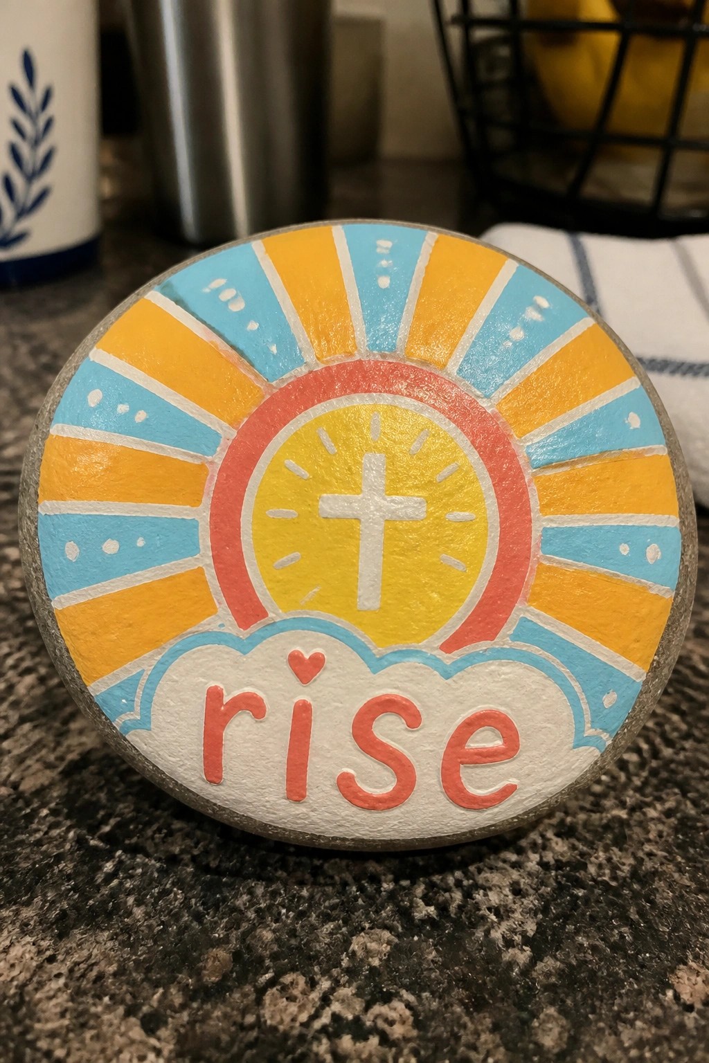 Colorful painted stone with sun rays, cross, clouds, and red word rise