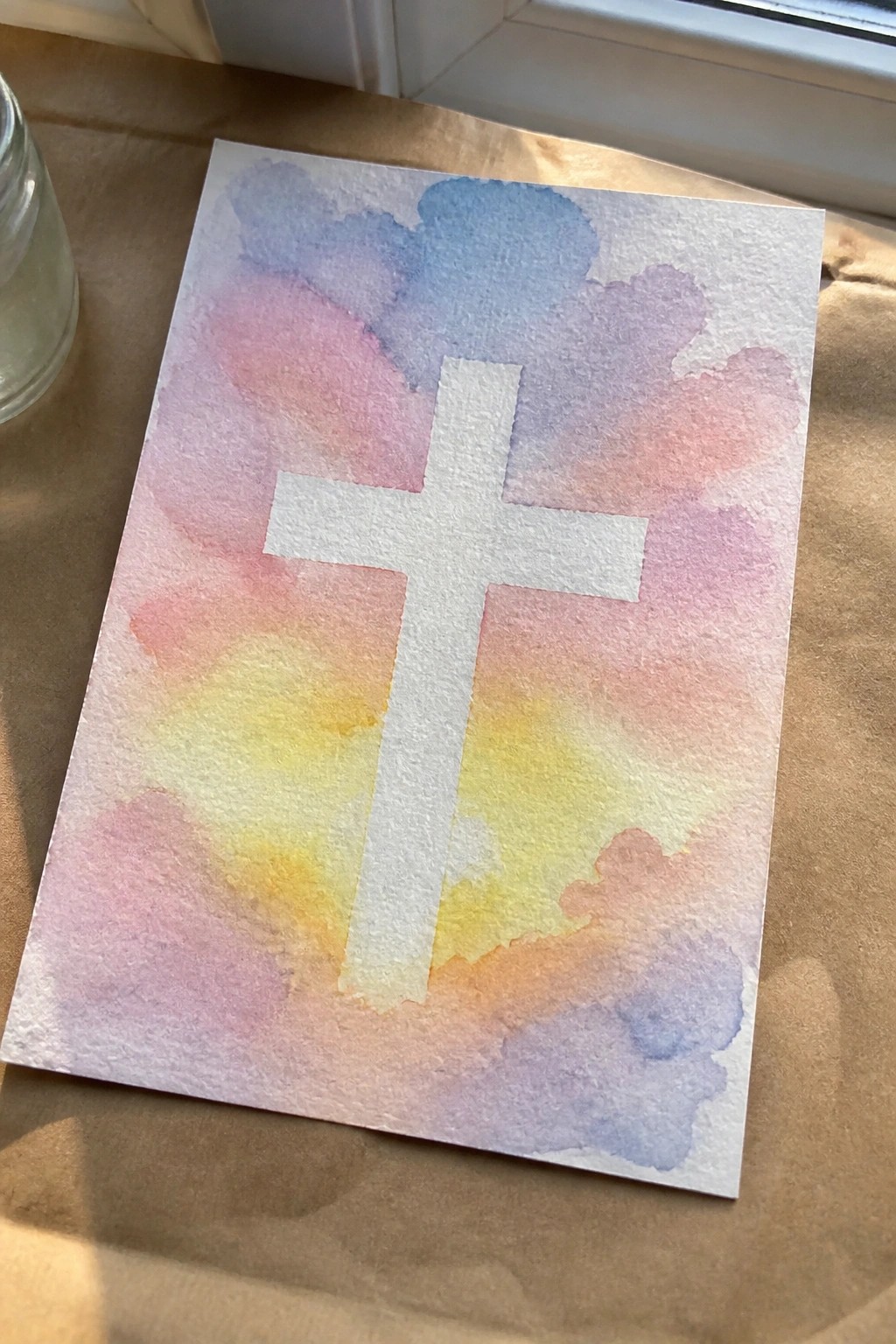 White cross centered on soft pastel watercolor clouds in pink, blue, and yellow.