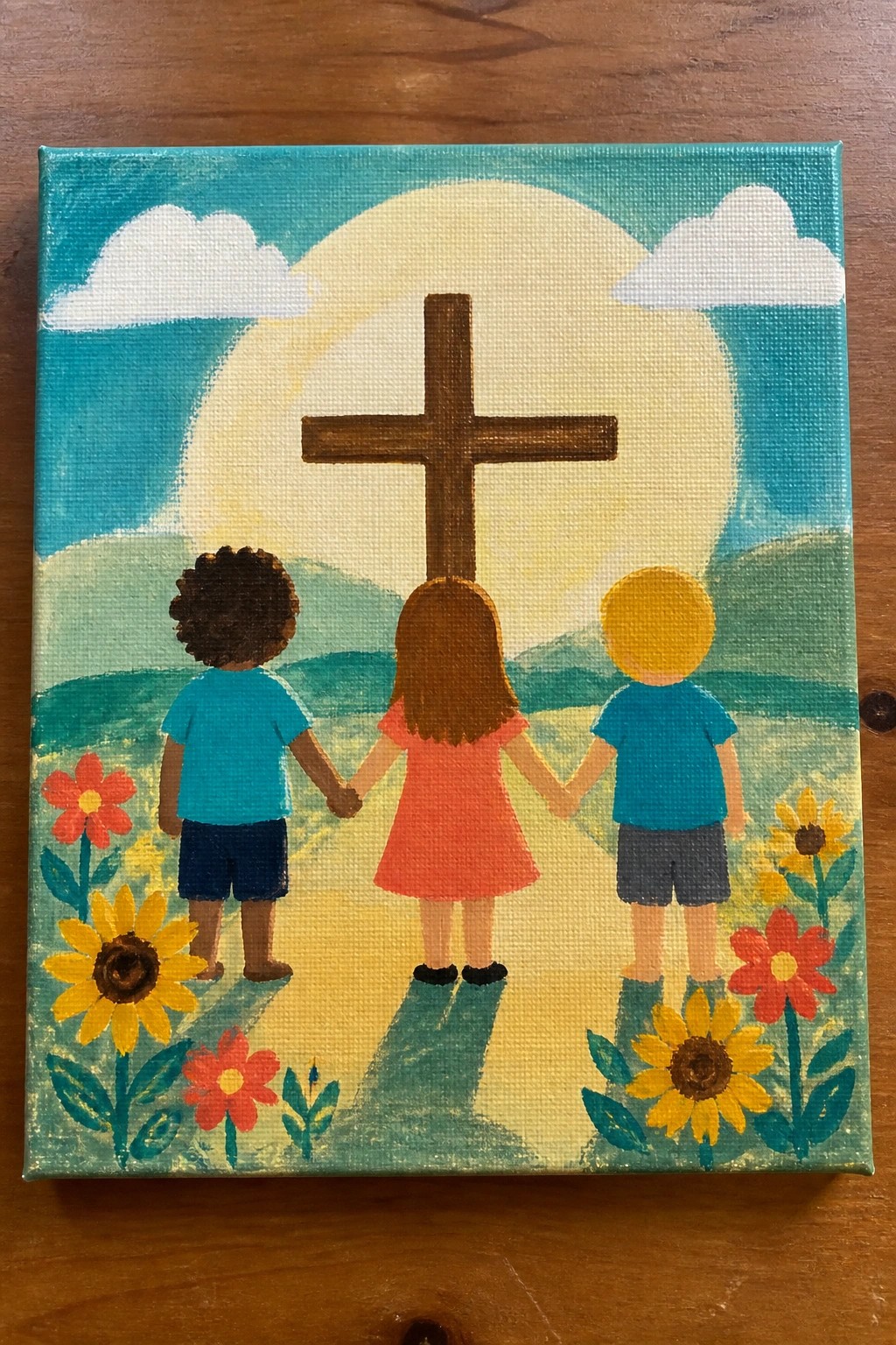 Three children holding hands before cross in sunny flower field with hills and clouds