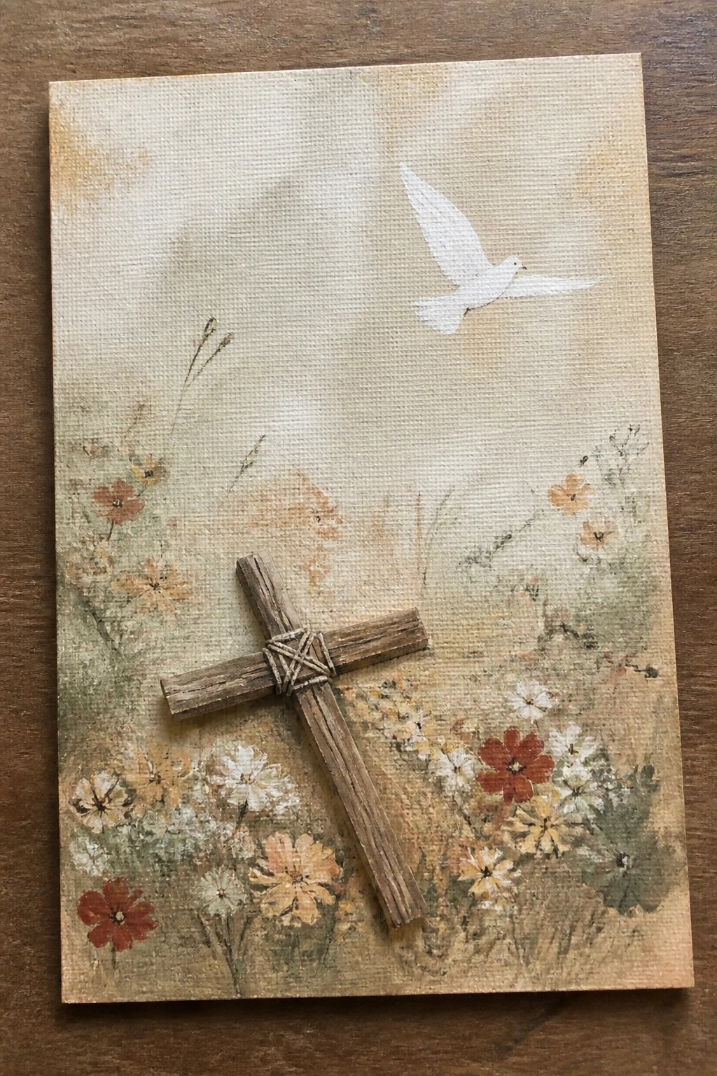 Twine-bound wooden cross with white dove above painted wildflowers on canvas.
