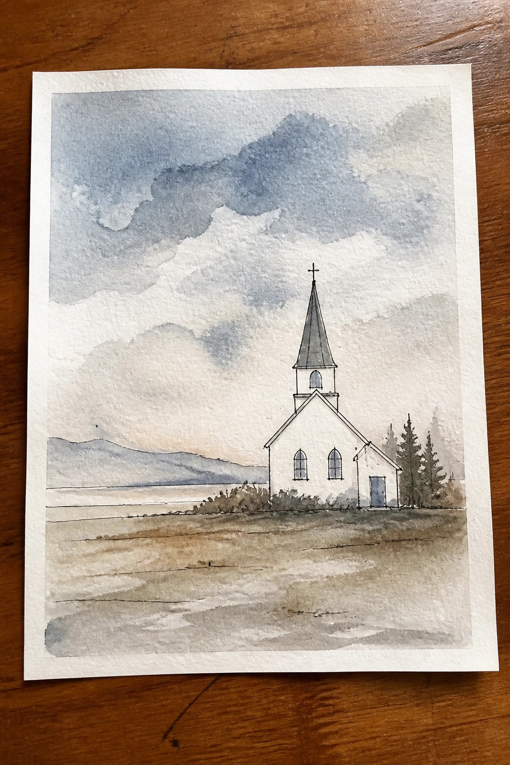 Watercolor of white church with steeple and cross in mountain landscape with fields