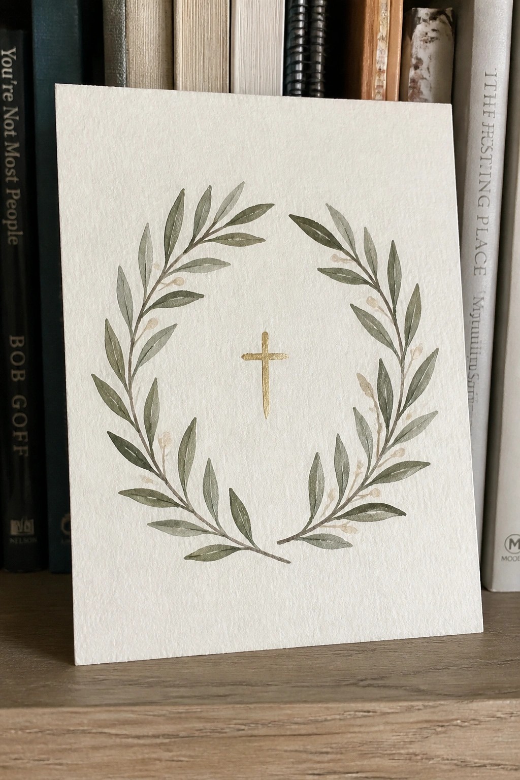 Watercolor white card with green leaf wreath and gold cross on wooden shelf