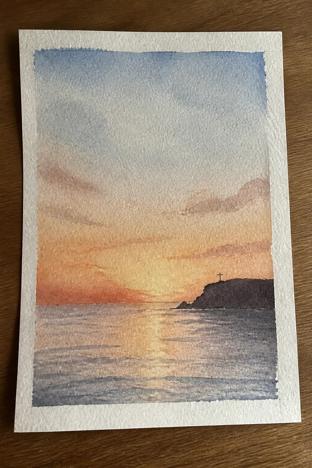 Watercolor sunset over calm sea with orange-pink sky, glowing reflections, and dark cliff with cross.