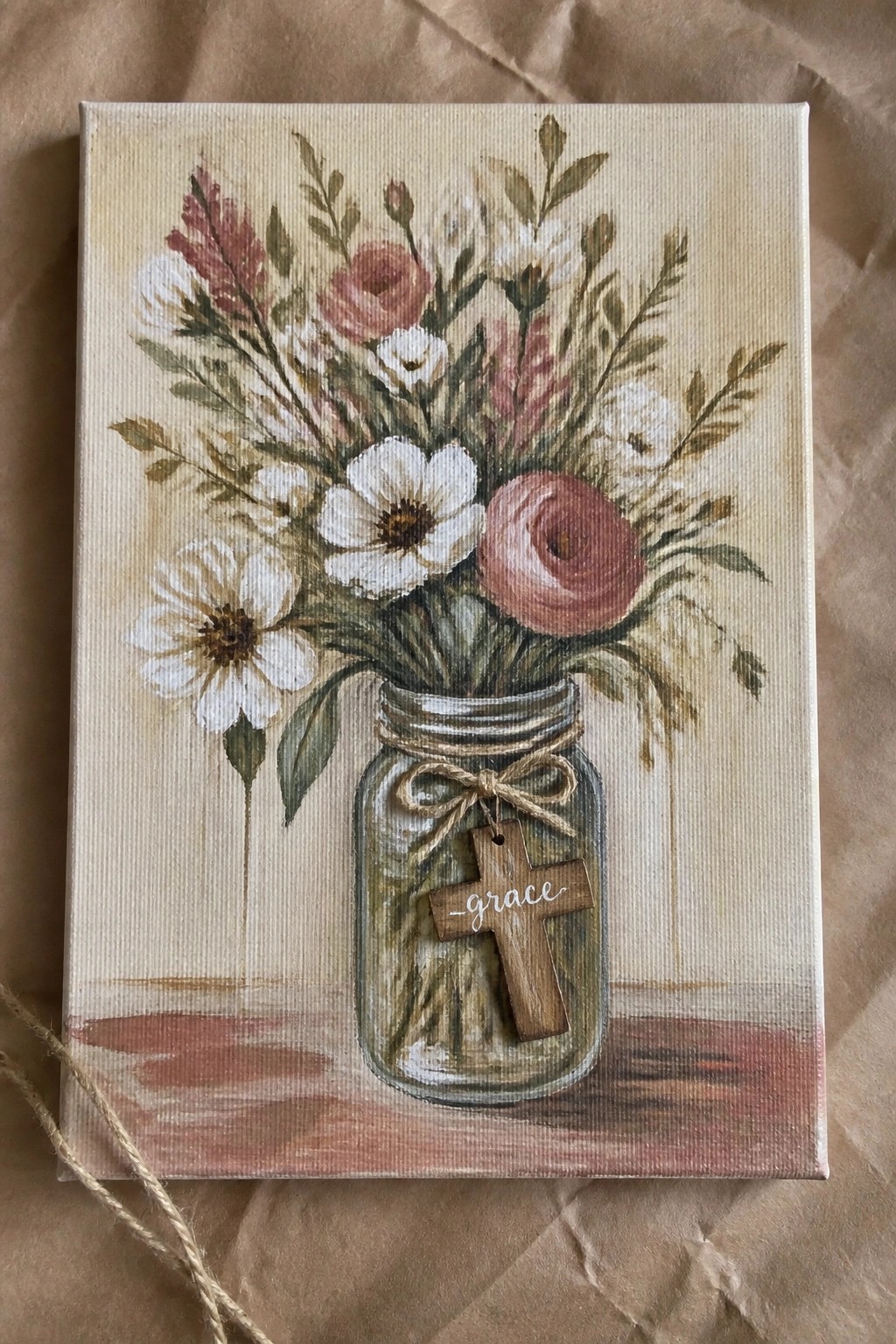 A canvas painting of pink and white flowers in a mason jar with a wooden cross labeled grace hanging from twine.