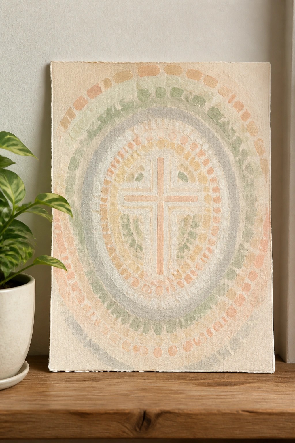 Pastel mosaic painting with central cross beside potted plant on wooden shelf