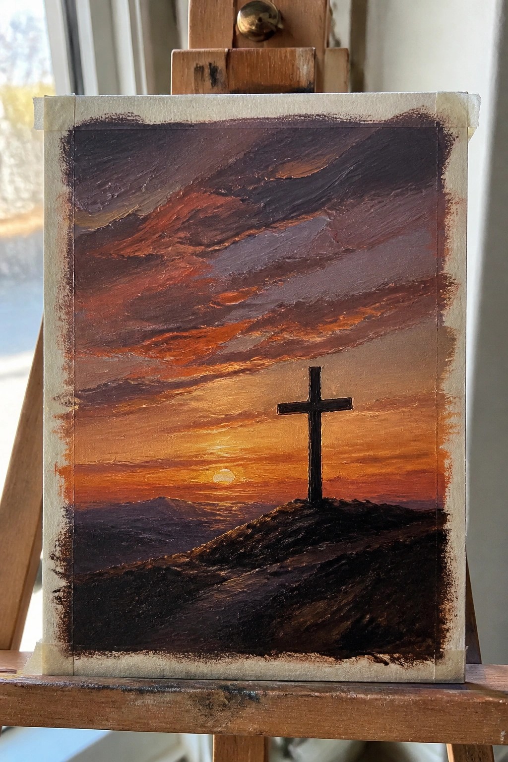 Black cross silhouetted on hill under vibrant red-orange sunset sky on easel.