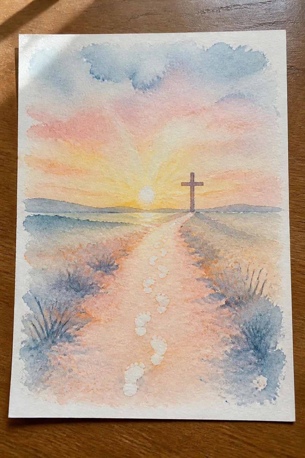 Watercolor painting of footprints on path leading to cross at colorful sunset
