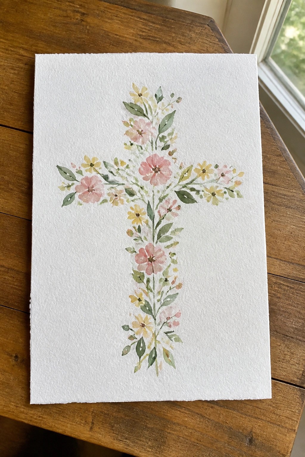 Watercolor floral cross with pink and yellow blooms on white paper atop wood table