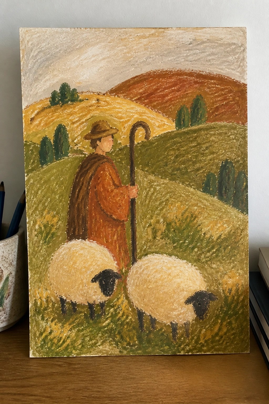 Shepherd in brown robe and hat with crook stands by two sheep in rolling hills.