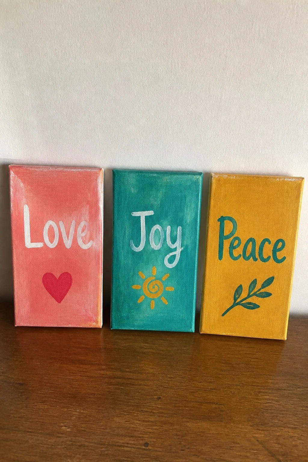 Three colorful canvases on wood: pink Love with heart, teal Joy with sun, yellow Peace with leaf.