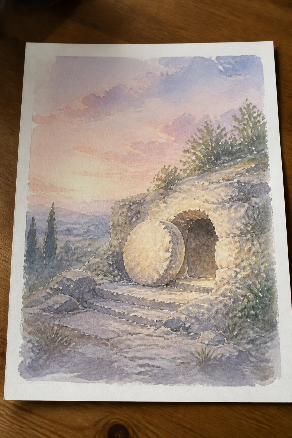 Watercolor of ancient stone tomb with large round boulder beside open entrance at sunset.