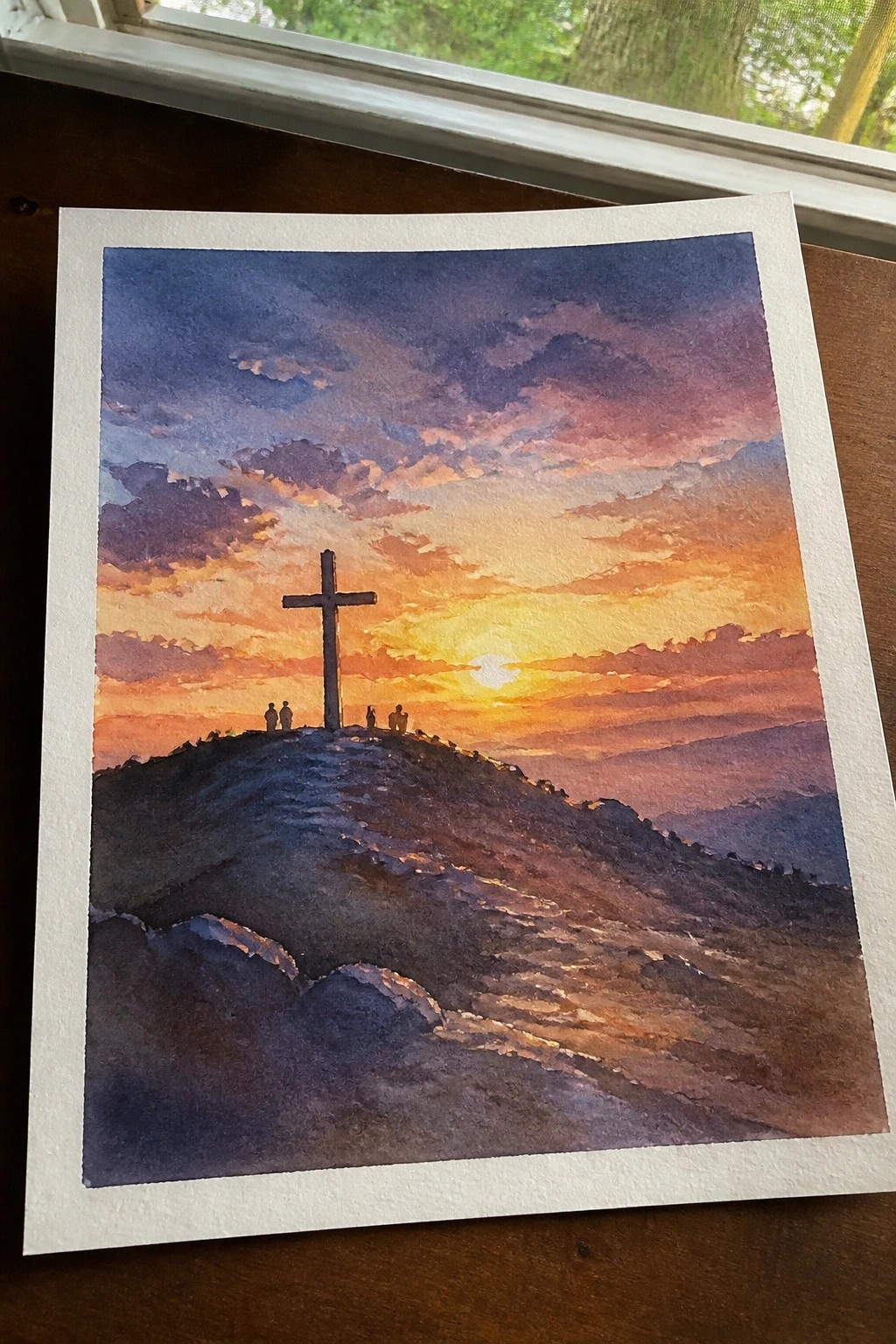 Watercolor painting of silhouetted cross on hill at vibrant orange-purple sunset with figures nearby.