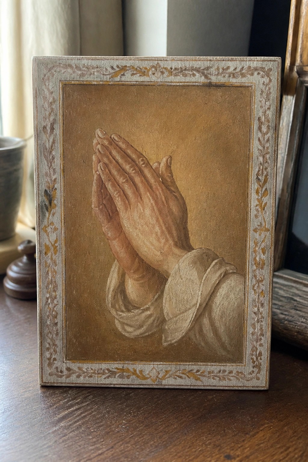 Painted praying hands with white sleeves in ornate leaf-patterned frame on wood.