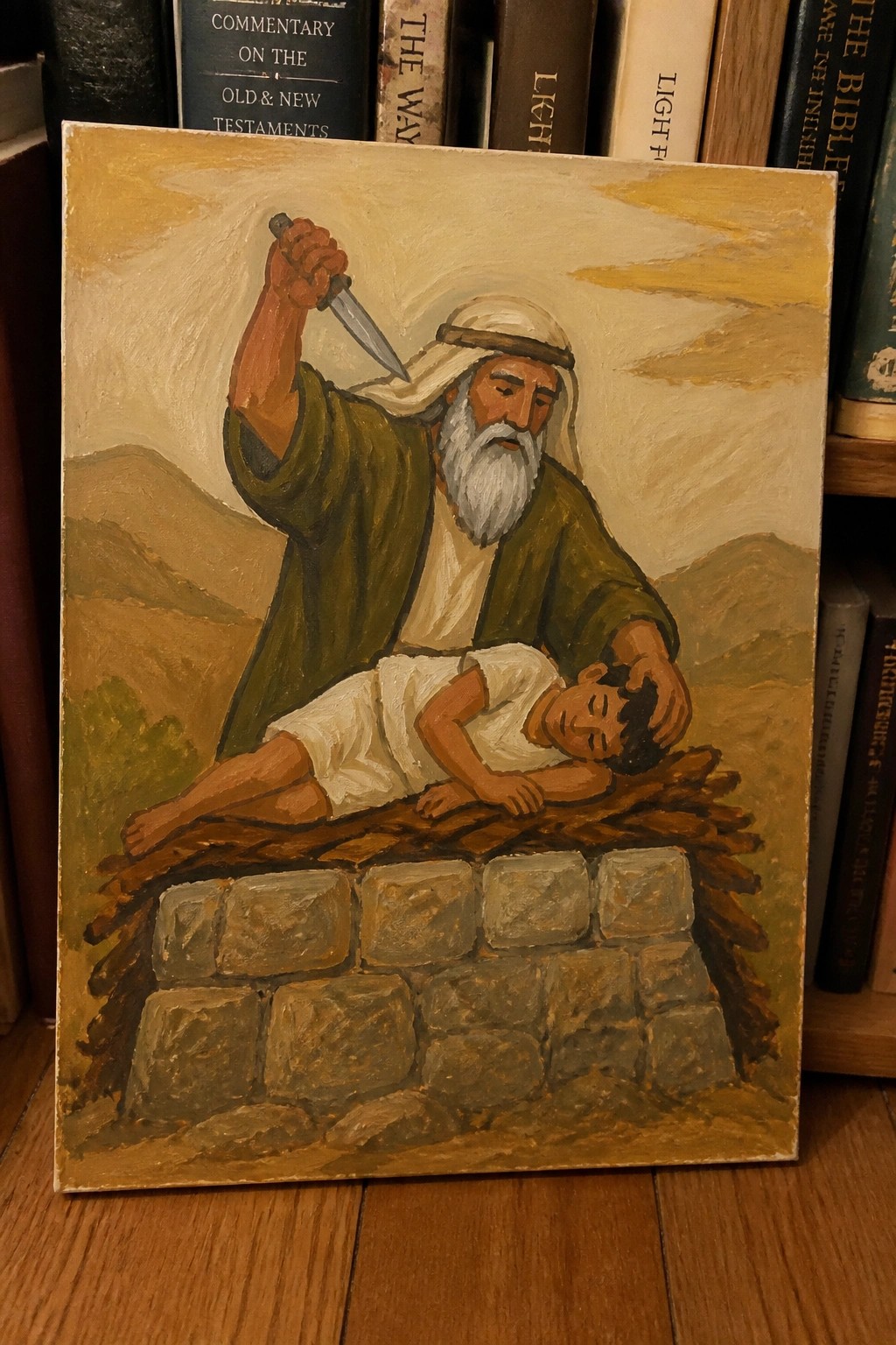 A painting shows a bearded man in green and white robes raising a knife above a boy lying on a wooden altar built on stones, with mountains behind them.