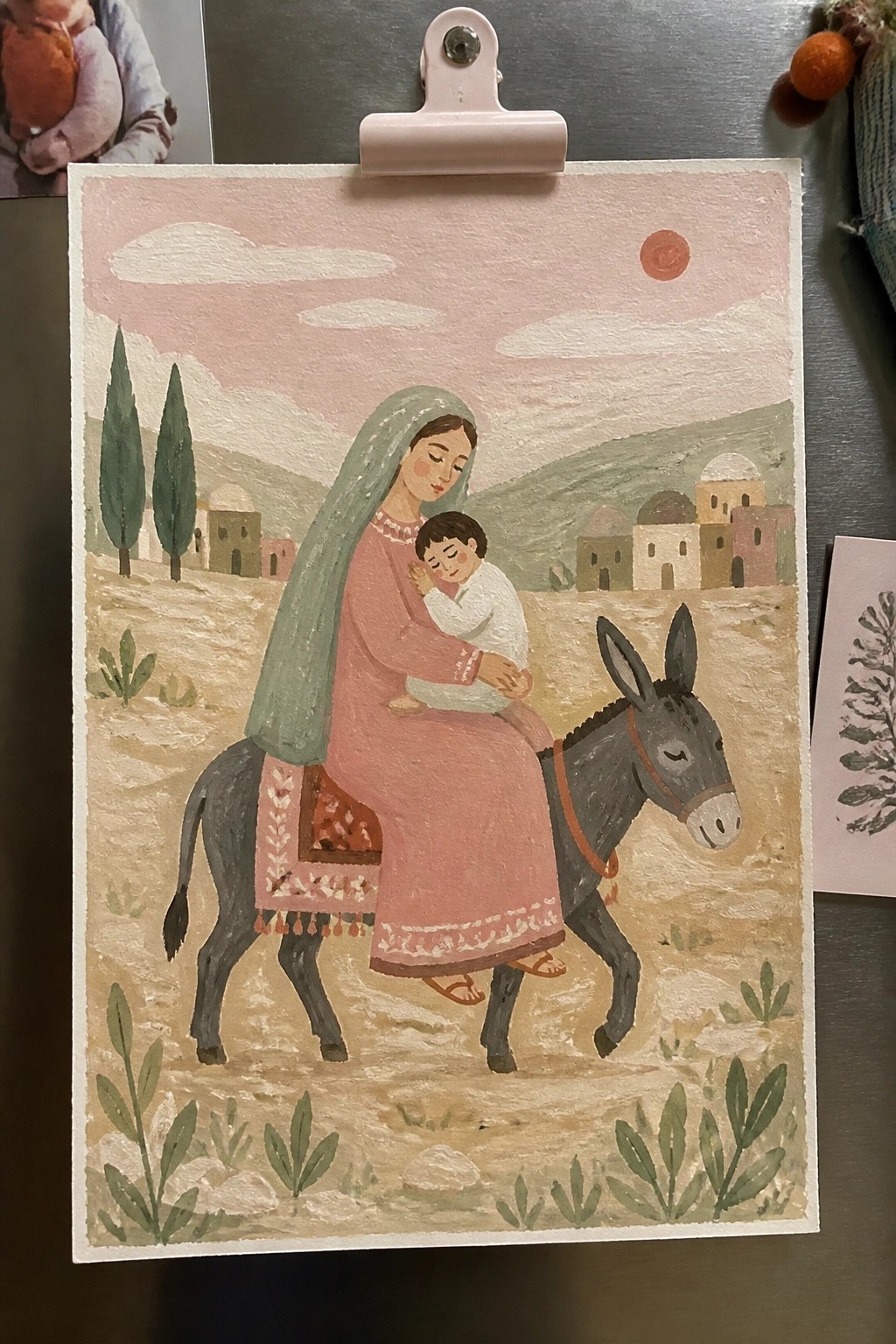Mother in pink dress with green veil cradles baby on gray donkey in pastel landscape