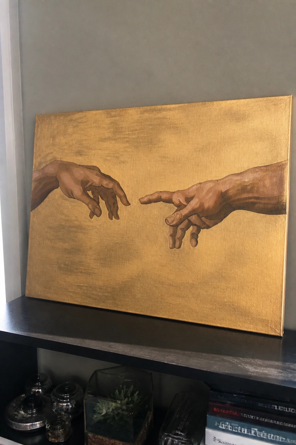 Gold canvas painting of two hands nearly touching, on black shelf with books