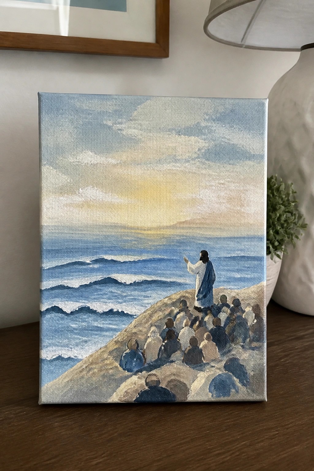 Robed Jesus raises hand before seated followers on hill overlooking ocean waves at sunset