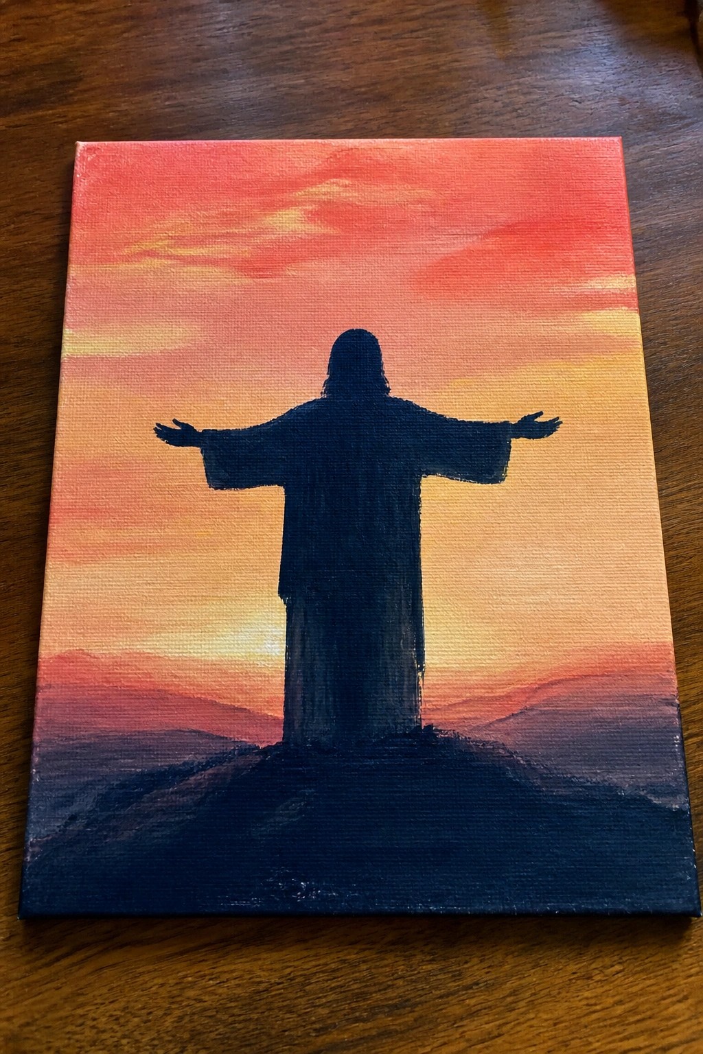 Silhouetted robed figure with arms outstretched atop hill under vibrant sunset sky.