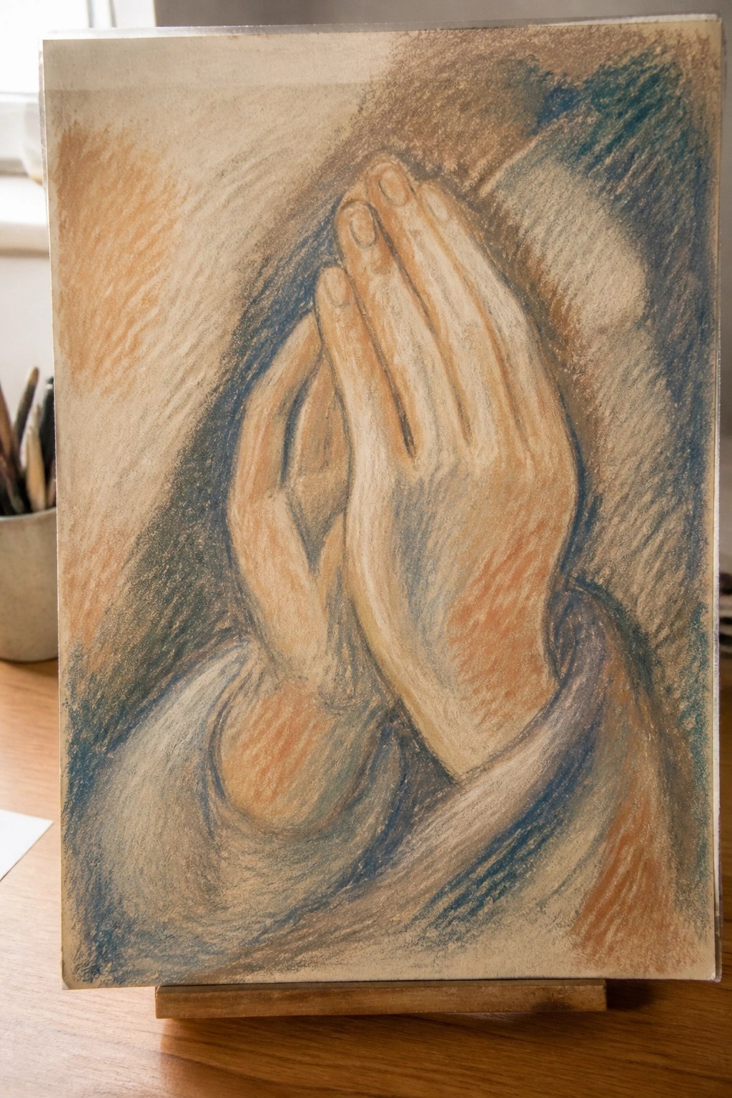 Pastel drawing of clasped praying hands in earthy blue and orange tones on easel