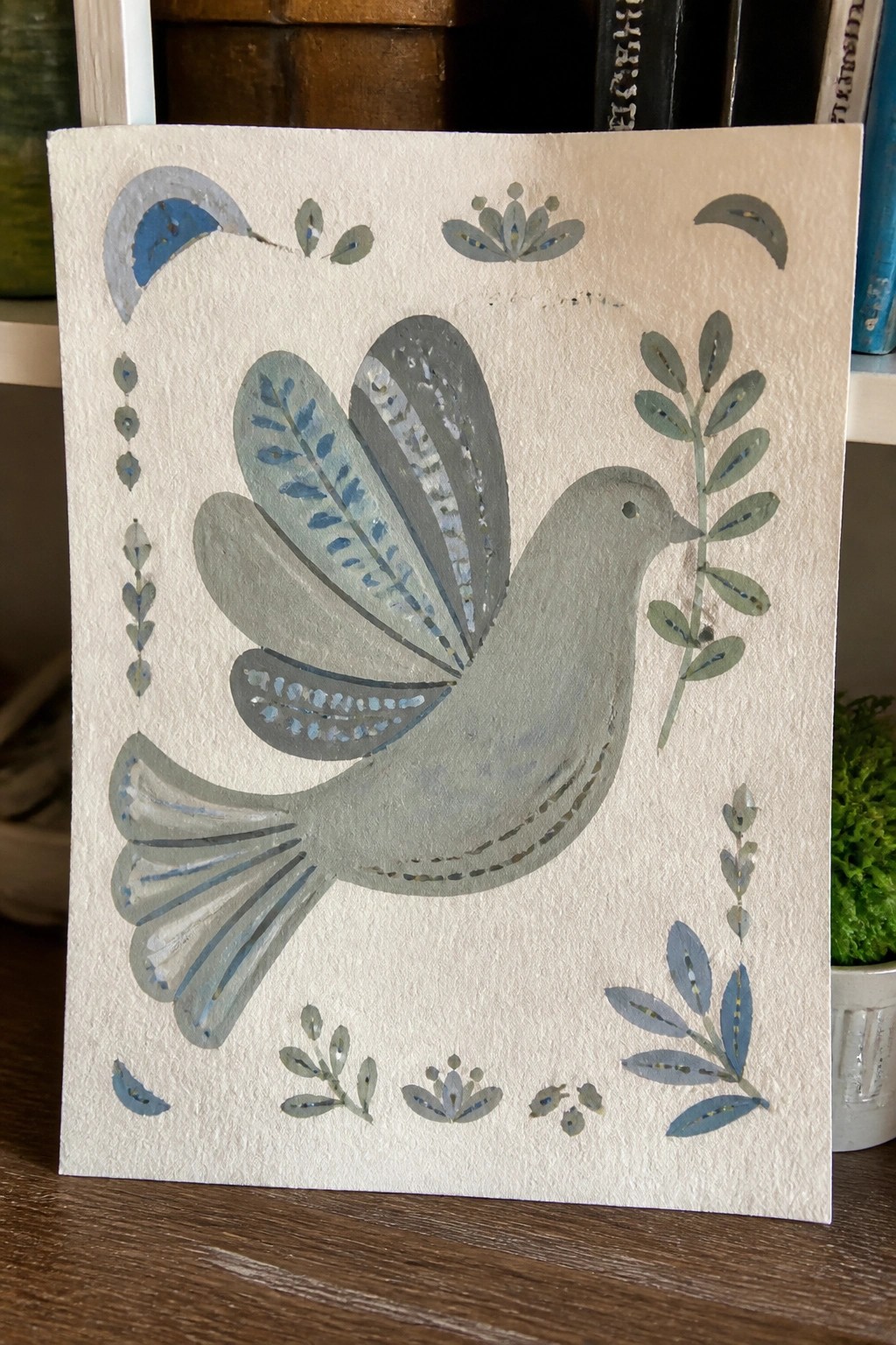Stylized gray dove with blue wing details holding a leafy branch amid decorative motifs.
