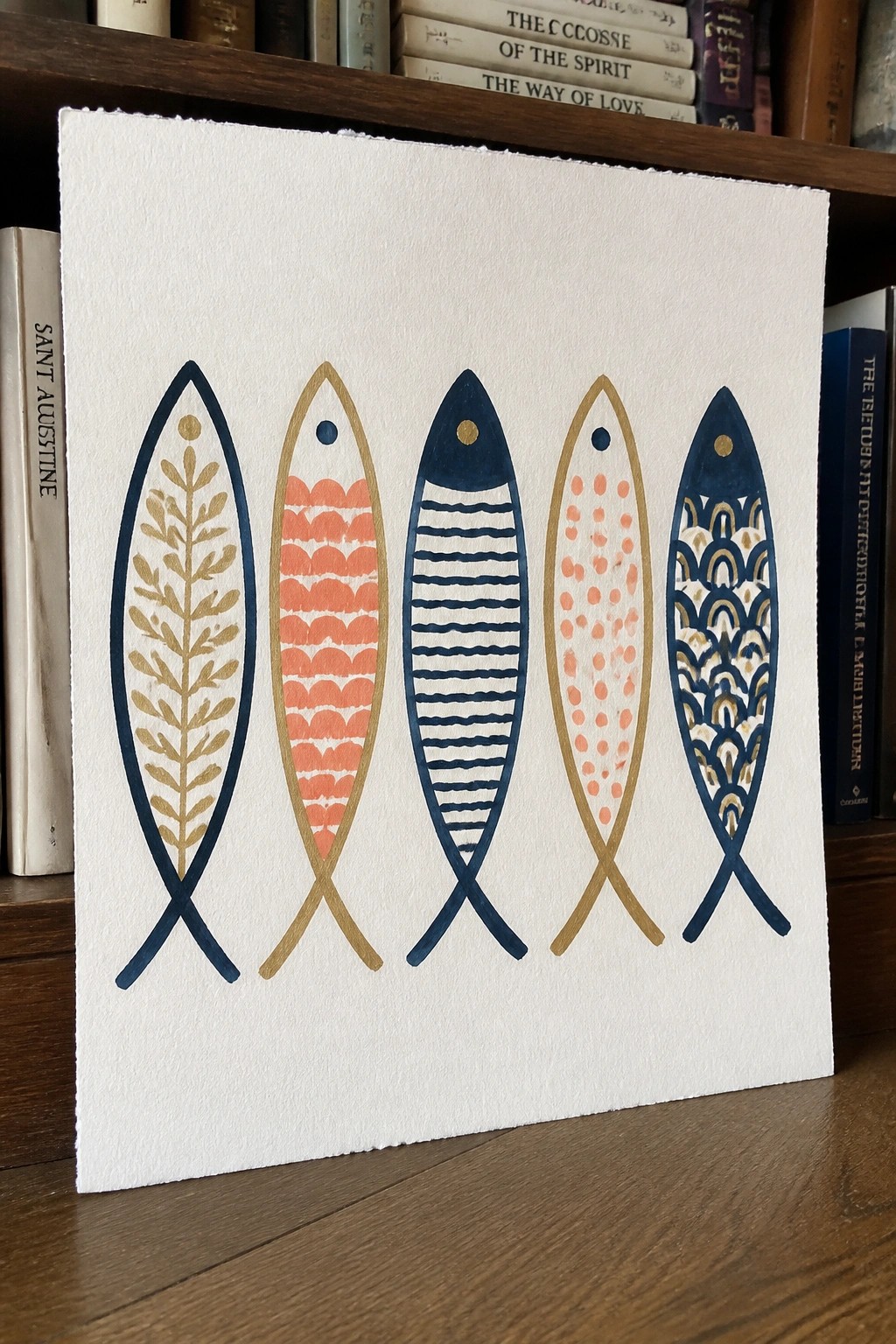 Five colorful patterned fish illustrations on white paper with books behind.