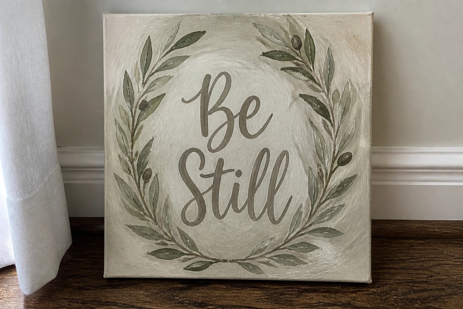 Beige canvas with green leaf wreath and "Be Still" text, leaning on wood floor