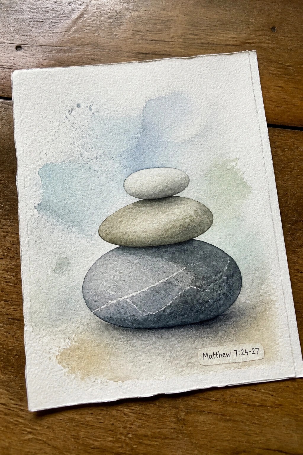 Three stacked stones in soft blue-green watercolor with Matthew 7:24-27 label