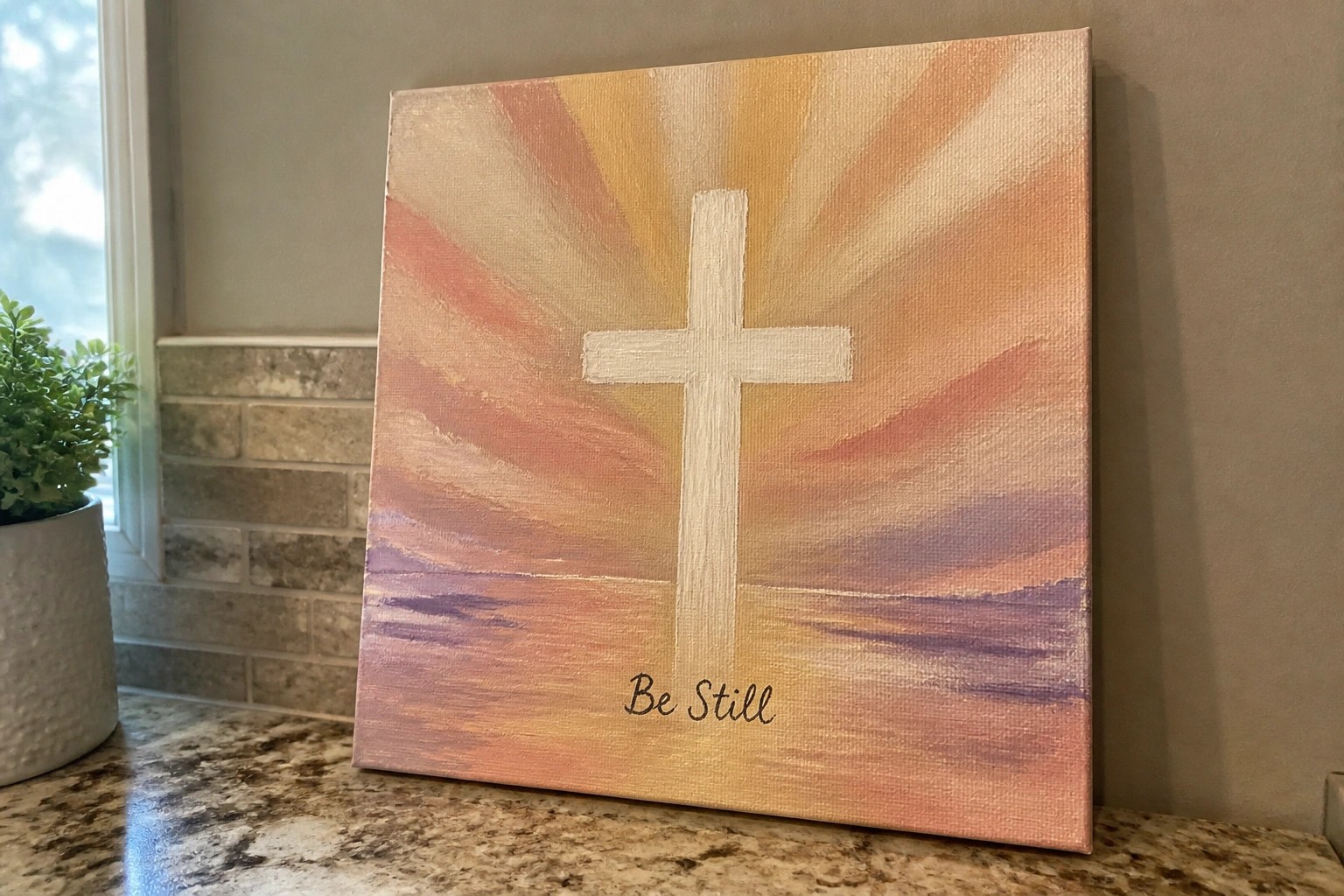 Canvas painting of white cross over orange-purple mountain sunset with "Be Still" text.
