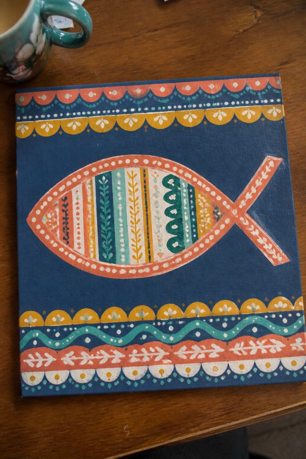 Colorful fish painting with patterned borders on blue canvas beside teal mug on wood