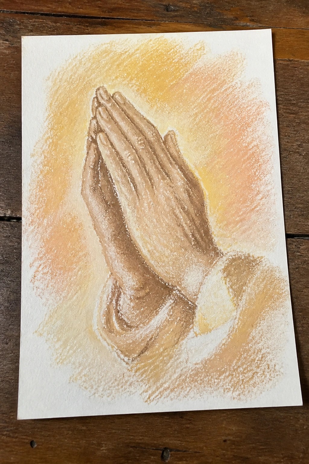 Clasped praying hands with brown skin and white sleeve against warm glowing pastel background.